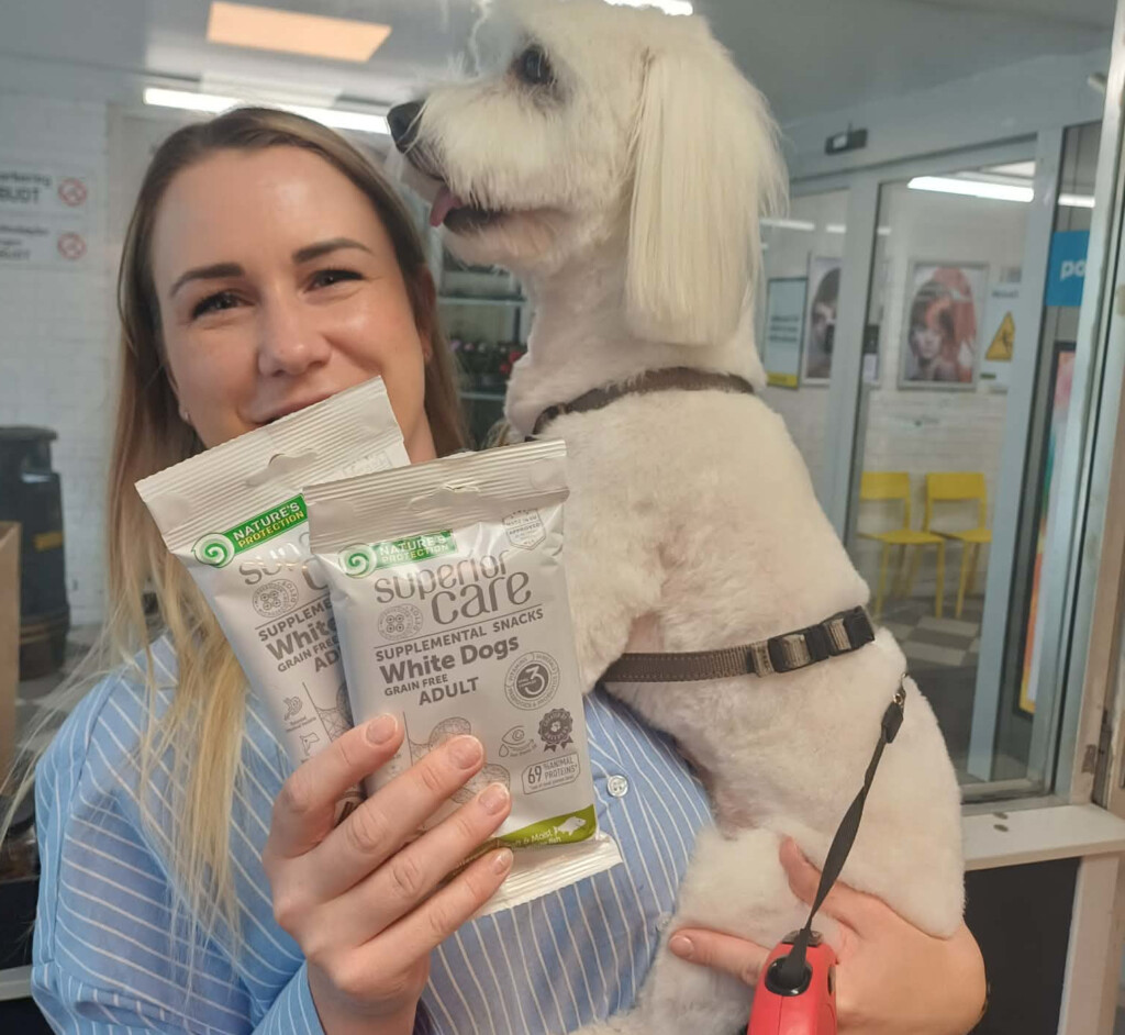 Ansatt i Hunddelux holder en hvit hund og to poser hundegodteri i salongen